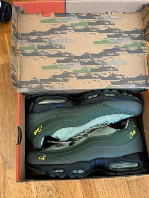 Nike Air Max 95 x Corteiz gröna sneakers - Nike Air Max 95 x Corteiz sneakers i olivgrönt och svart med gula detaljer. Skorna har synliga Air-enheter i sulan, mesh och läderpaneler samt Corteiz-logga broderad på sidan. Snygg box med unik design medföljer.