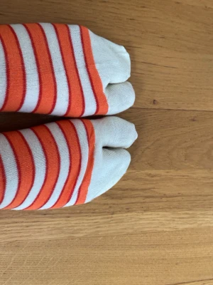 Randiga tåstrumpor i orange och vitt - Säljer ett par tåstrumpor med orange och vita ränder. Strumporna har separata fack för varje tå och är tillverkade i mjuk bomull. Perfekta för dig som vill ha något annorlunda och bekvämt på fötterna.