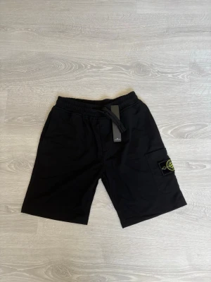 Svarta shorts från Stone Island - Säljer ett par svarta shorts från Stone Island med elastisk midja och snörning. Shortsen har en ficka på sidan med den klassiska Stone Island-loggan och är tillverkade i mjuk bomull. Perfekta för en avslappnad och stilren look.