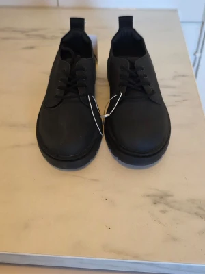 Svarta loafers med grov sula - Stilrena svarta loafers med snörning och grov sula. Skorna har en klassisk rund tå och är tillverkade i ett matt material som ger en modern look. Perfekta för dig som vill ha en enkel men trendig sko till vardagslooken.