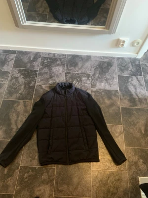 Svart pufferjacka med dragkedja - Snygg svart pufferjacka med ståkrage och hel dragkedja framtill. Jackan har två sidofickor och quiltad design på framsidan. Ärmarna är släta och materialet ser ut att vara en blandning av polyester och syntet. Perfekt för kalla vinterdagar.