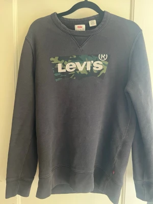 Svart sweatshirt från Levi's med camologga - Cool svart sweatshirt från Levi's med klassisk logga i vitt och camomönster på bröstet. Tröjan har rund halsringning, ribbade muddar och är tillverkad i mjuk bomullsblandning. Perfekt för en avslappnad streetwear-look. Passar om du är runt 170