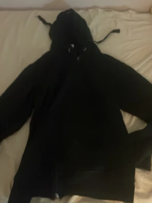 Svart hoodie med dragkedja - En klassisk svart hoodie med dragkedja framtill och justerbar huva med snören. Hoodien har långa ärmar och ribbade muddar vid ärmslut och nederkant. Perfekt för en avslappnad och stilren look.