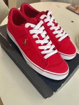 Röda Polo Ralph Lauren sneakers - Snygga röda sneakers från Polo Ralph Lauren med vit sula och vita skosnören. Skorna har klassisk låg modell och är tillverkade i canvasmaterial. Ikoniska Polo-loggan broderad på sidan. Perfekta för en sportig och trendig look. Använd 1 gång 