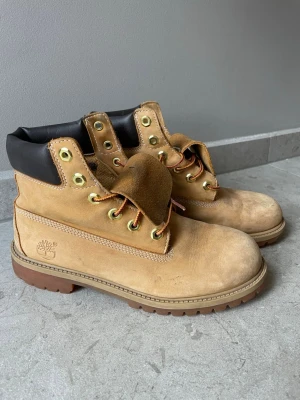 Timberland kängor i nubuck strl 39 - Klassiska beige kängor från Timberland i slitstark nubuck med robust gummisula och vadderad svart krage. Guldiga metalldetaljer och gulbruna snören ger en snygg kontrast. Perfekta för höst och vinter när du vill ha både stil och funktion. Strl 39