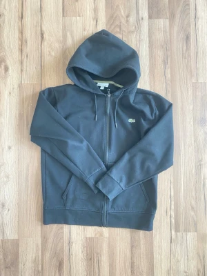 Svart hoodie från Lacoste med dragkedja - Säljer en svart hoodie från Lacoste i klassisk passform. Hoodien har dragkedja framtill, två fickor och den ikoniska gröna krokodilloggan på bröstet. Tillverkad i mjukt bomullsmaterial och har justerbar huva med snören.