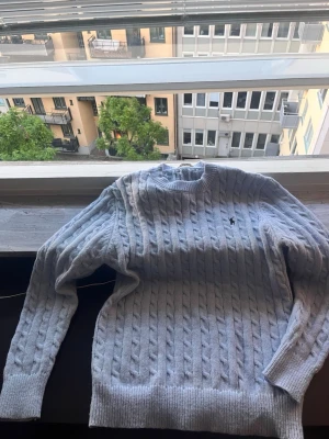 Ljusblå kabelstickad tröja Polo Ralph Lauren - Ljusblå kabelstickad tröja från Polo Ralph Lauren med klassisk rund halsringning och broderad logga på bröstet. Tröjan har ribbade muddar vid ärmslut och nederkant. Perfekt för lager-på-lager och snygg till jeans.