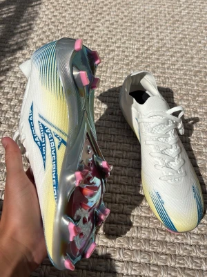 Adidas X Speedflow fotbollsskor vita - Säljer ett par Adidas X Speedflow fotbollsskor i vitt med blå och gula detaljer. Skorna har en lätt ovandel i syntetmaterial och en skinande sula med rosa dobbar. Perfekta för dig som vill ha snabbhet och stil på planen.