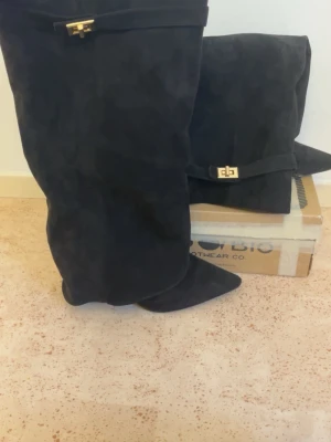 Svarta overknee stövlar med gulddetalj - Säljer ett par svarta overknee stövlar i mockaimitation med spetsig tå och dekorativ guldfärgad spänndetalj upptill. Stövlarna har en slouchy passform och är perfekta för att lyfta vilken outfit som helst.