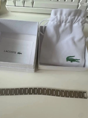 Lacoste armband - Snyggt armband från Lacoste i silverfärgat rostfritt stål med stilrena, rektangulära länkar. Armbandet har en diskret Lacoste-logga ingraverad på spännet och levereras med original ask och tygpåse. Perfekt accessoar för en modern look.