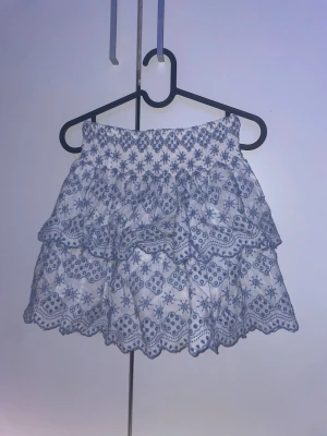 Vit och blå volangkjol från Lindex - Supersöt kort kjol från Lindex i vitt med blått blommigt broderi och volanger. Kjolen har resår i midjan och vågig kant nertill. Perfekt för dig som gillar romantisk och somrig stil.
