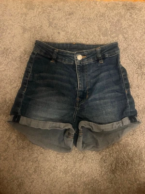 Mörkblå denimshorts med uppvik - Snygga mörkblå jeansshorts med hög midja och uppvikta benslut. Klassisk femficksmodell med knapp och dragkedja framtill. Perfekta för sommaren och lätta att matcha med olika toppar.
