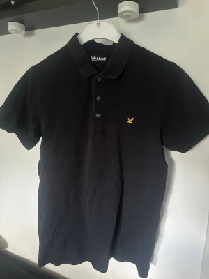 Svart piké från Lyle & Scott - Klassisk svart pikétröja från Lyle & Scott med gul broderad logga på bröstet. Tröjan har korta ärmar, krage och knappar framtill. Tillverkad i mjuk bomull som känns skön mot huden.