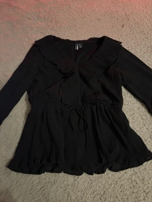 Svart blus med volangkrage och knyt - Säljer en svart blus från Vero Moda med långa ärmar, volangkrage och knyt i midjan. Blusen har en lätt rynkad struktur och volangdetaljer vid ärmslut och nederkant. Perfekt för dig som gillar en romantisk och trendig stil.