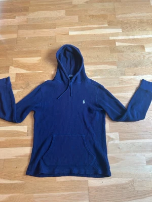 Marinblå hoodie från Polo Ralph Lauren - Säljer en marinblå hoodie från Polo Ralph Lauren med huva och dragsko. Tröjan har en klassisk känguruficka framtill och det ikoniska Polo-märket broderat på bröstet. Tillverkad i ett ribbat, mjukt material som känns skönt mot huden. Perfekt för en avslappnad stil.