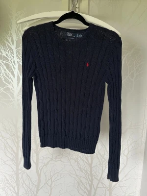 Mörkblå kabelstickad tröja Polo Ralph Lauren - Mörkblå kabelstickad tröja från Polo Ralph Lauren.. Klassisk rund halsringning och lång ärm. Liten röd broderad logga på bröstet. Perfekt till jeans eller kjol för en stilren look.