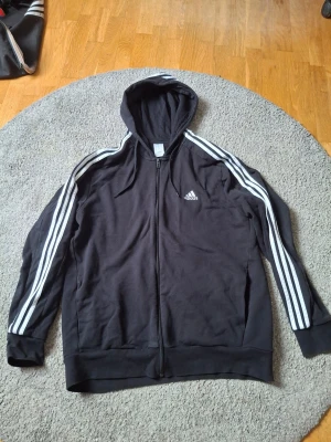 Svart Adidas hoodie med dragkedja XL - Säljer en svart hoodie från Adidas med dragkedja och huva. Klassiska vita ränder längs ärmarna och Adidas-logga på bröstet. Tillverkad i mjukt material, perfekt för en sportig och avslappnad stil. Långa ärmar och ribbade muddar.