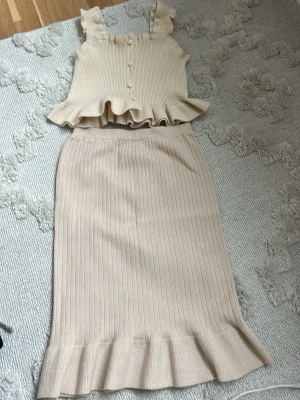 Beige ribbat set med volangdetaljer - Säljer ett beige ribbat set med linne och kjol. Linnet har breda axelband, volangkant nedtill och dekorativa knappar framtill. Kjolen är midilång med ribbad struktur och volangkant längst ner. Perfekt för en chic och avslappnad look.