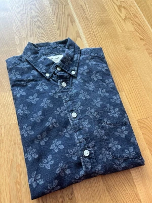 Blommig blå skjorta från East West - Snygg blå skjorta från East West med blommigt mönster och vita knappar. Skjortan har klassisk krage och bröstficka, perfekt för en avslappnad men ändå stilren look. Tillverkad i mjuk bomull som känns skön mot huden. Kortärmad 