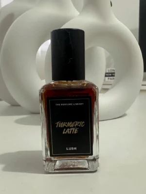 Lush Turmeric Latte parfym 30ml - Unik parfym från Lush, Turmeric Latte, i en rektangulär glasflaska med svart etikett och svart lock. Doften är inspirerad av gurkmeja och latte, vilket ger en varm och kryddig känsla. 