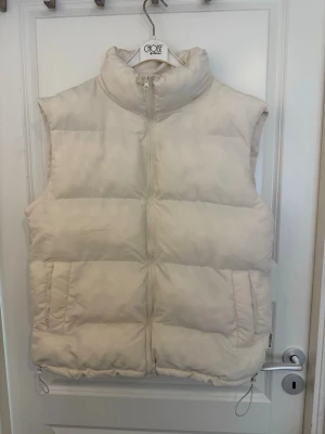 Vit dunväst med hög krage - Stilren vit dunväst med hög krage och dragkedja framtill. Västen har två fickor på sidorna och justerbar dragsko nedtill. Perfekt för lager-på-lager och ger en clean look till din outfit. Passar dig som gillar minimalistisk och trendig stil. Sömmarna sönder i armhålan men inget som syns vid användning. 