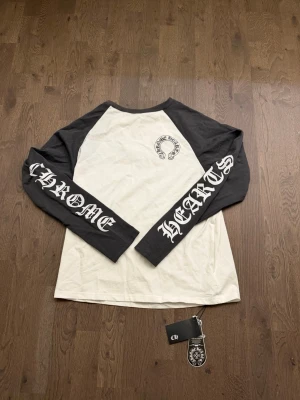 Long sleeve tröja - Cool långärmad topp med svartvita kontraster och tryck på både ärmar och rygg. Raglanärmar i mörkgrått och kropp i vitt, med tydliga logotyper och gotisk text. Perfekt för dig som gillar streetwear och unika detaljer.