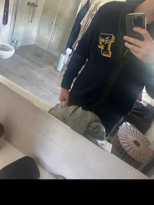 Mörkblå collegekofta med grön kant - Säljer en mörkblå stickad collegekofta med gröna ribbstickade kanter och knappar framtill. På bröstet finns ett gult och vitt broderat märke med bokstaven 'L' och ett vingskor-motiv. Perfekt för dig som gillar preppy stil.