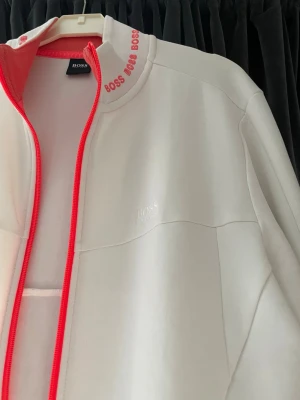 Vit zip hoodie från BOSS - Snygg vit hoodie från BOSS med dragkedja och röda detaljer längs dragkedjan och i kragen. BOSS-logga broderad på bröstet och tryckt text i kragen. Tillverkad i mjukt material, perfekt för en clean och sportig look.