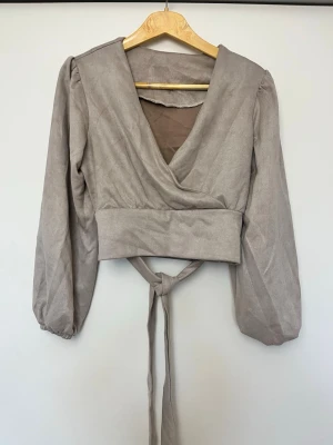 Beige omlottopp med knytband - Trendig beige omlottopp med långa puffärmar och bred midjeresår. Toppen har knytband i midjan och djup v-ringning framtill. Perfekt att styla med höga jeans eller kjol för en snygg look.
