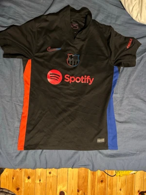 Svart FC Barcelona matchtröja Nike - Snygg svart FC Barcelona matchtröja från Nike med röda och blå detaljer på sidorna. Tryck med Spotify-logga på bröstet och UNHCR-logga bak. Nummer 10 och namnet Alejandro på ryggen. Korta ärmar och sportig passform, perfekt för fotboll eller träning.