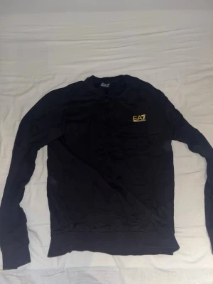 Svart sweatshirt från EA7 Emporio Armani - Svart långärmad sweatshirt från EA7 Emporio Armani med guld logga på bröstet. Klassisk rund halsringning och ribbade muddar vid ärmslut och nederkant. Perfekt för en avslappnad och stilren look.