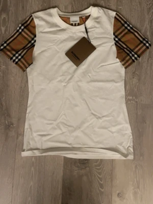 Vit Burberry t-shirt med rutiga ärmar - Snygg vit t-shirt från Burberry med klassiska beige, bruna och svarta rutiga kortärmade ärmar. Tillverkad i mjuk bomull och har en normal passform. Perfekt för dig som vill ha en stilren look med en twist. Är storlek S men sitter som XS