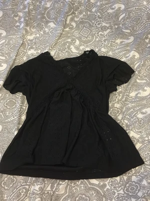 Svart blus med broderade detaljer - Svart blus med korta ärmar och vacker broderad spets längs framstycket och halsen. Blusen har en luftig passform och små hålmönster som ger en cool look. Perfekt för dig som gillar stilrena plagg med lite extra detaljer.
