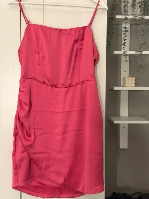 Rosa satin klänning med smala axelband - Snygg rosa kort klänning i satin med smala axelband och lätt drapering på sidan. Klänningen har en figurnära passform och är perfekt för dig som vill sticka ut med en färgstark look.