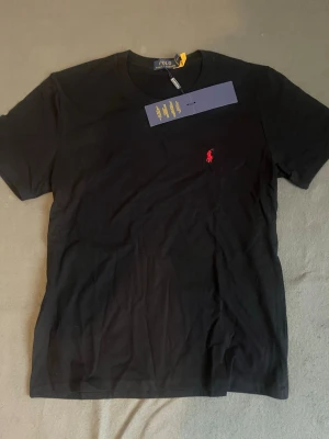 Svart t-shirt från Polo Ralph Lauren - Säljer en svart t-shirt från Polo Ralph Lauren med klassisk röd logga broderad på bröstet. T-shirten har rund hals och korta ärmar, perfekt för en clean och stilren look. Materialet är mjuk bomull som känns skönt mot huden.