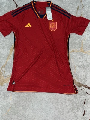 Spaniens 2022 player kit  - Snygg röd fotbollströja från Adidas med Spaniens landslagsmärke på bröstet och gula detaljer. Tröjan har mörkblå inslag på axlar och sidor samt klassiska Adidas-ränder. Tillverkad i lätt polyester, perfekt för träning eller match. Storlek M men passar även S om man vill ha lösare passform 