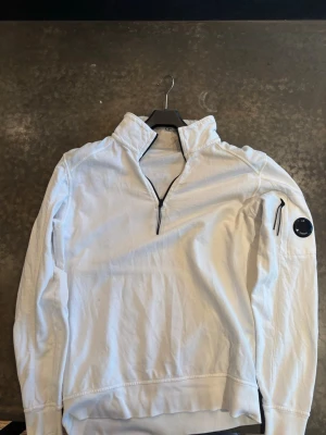 Vit half-zip tröja från C.P. Company - Snygg vit half-zip tröja från C.P. Company med hög krage och svart dragkedja. Tröjan har en ikonisk linsdetalj på ärmen och en liten ficka med dragkedja. Perfekt för en clean och modern stil.