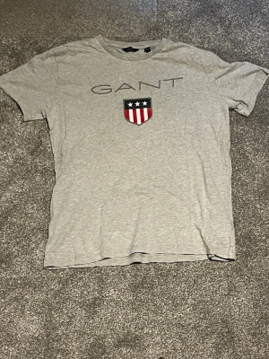 Grå GANT t-shirt med logga - Snygg grå t-shirt från GANT med klassisk rund hals och tryckt logga på bröstet. På framsidan finns ett stort GANT-tryck och en sköld med stjärnor och ränder i rött, vitt och blått. Perfekt till jeans eller shorts och speciellt nu till sommaren. denna t-shirt passar alla som är mellan 160 cm lång till 180 cm lång eftersom passformen är stretchig och bekväm. Vid minsta fråga eller fundering var vänligen att meddela mig. Med vänliga hälsningar Eric 
