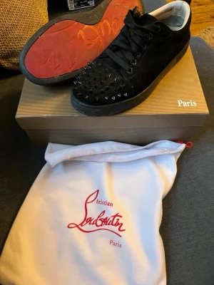 Svarta Christian Louboutin sneakers - Säljer ett par svarta sneakers från Christian Louboutin med klassisk röd sula och svarta nitar på tån. Skorna är låga och har ovandel i mocka, svarta skosnören och en vit insida. Kommer med originalpåse och låda.