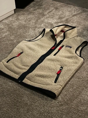 Beige fleeceväst från Patagonia - Säljer en beige fleeceväst från Patagonia med svarta och röda detaljer. Västen har tre fickor med dragkedja, hög krage och svarta kantband. Materialet är mjukt och fluffigt, perfekt för lager-på-lager. Klassisk Patagonia-logga på bröstet. Västen är knappt använd och är 40cm axel bred och 70cm lång