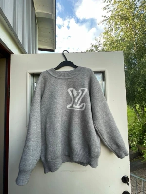 Grå Louis Vuitton tröja - Mysig grå tröja från Louis Vuitton med stor vit LV-logga på bröstet. Tröjan har rund halsringning, ribbade muddar och är tillverkad i ett mjukt, stickat material. Perfekt för dig som gillar stilrena och exklusiva plagg.