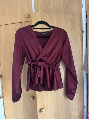 Vinröd omlottblus från Missay - Snygg vinröd omlottblus från Missay med lång ärm och knytband i midjan. Blusen har en djup V-ringning och ett lätt glansigt tyg som ger en elegant look. Perfekt att styla med jeans eller kjol för en trendig outfit.