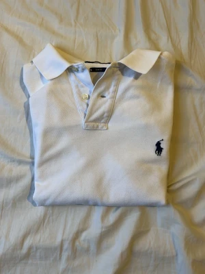 Vit pikétröja från Polo Ralph Lauren - Klassisk vit pikétröja från Polo Ralph Lauren med blå logga broderad på bröstet. Tröjan har korta ärmar, krage och knappar framtill. Tillverkad i mjuk bomull som känns skön mot huden. Perfekt till jeans eller shorts för en clean look.