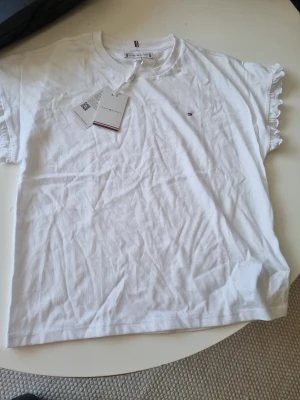 Vit t-shirt med volang från Tommy Hilfiger - Snygg vit t-shirt från Tommy Hilfiger med liten logga på bröstet och volangdetaljer vid ärmsluten. Klassisk rund halsringning och korta ärmar. Tillverkad i mjuk bomull som känns skön mot huden. Perfekt till jeans eller kjol för en clean look. Aldrig andvänts.