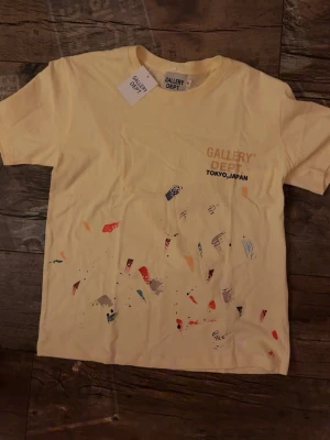 Gul t-shirt från Gallery Dept - Säljer en gul t-shirt från Gallery Dept med trycket 'GALLERY DEPT TOKYO, JAPAN' på bröstet och färgstänk i olika nyanser framtill. T-shirten har korta ärmar och en klassisk passform. Perfekt för dig som gillar streetwear och unika detaljer.