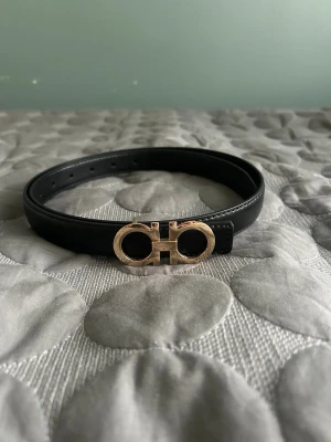 Svart skärp från Ferragamo i skinn - Snyggt svart skärp från Ferragamo i äkta skinn med ikoniskt guldfärgat dubbel-Gancio-spänne. Perfekt accessoar för att lyfta din outfit med en lyxig touch. Justerbar längd och klassisk design.
