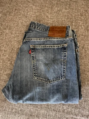Blå Levi's raka jeans - Säljer ett par klassiska blå Levi's jeans med rak passform och fem fickor. Jeansen har den ikoniska röda Levi's-lappen på bakfickan och en brun läderpatch i midjan. Tillverkade i slitstark denim, perfekta för en avslappnad stil.