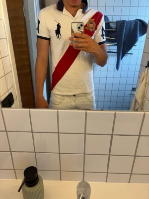 Vit pikétröja från Ralph Lauren - Säljer en vit pikétröja från Ralph Lauren med stor marinblå logga på bröstet, röd diagonal rand och broderad patch. Nummer 4 på ärmen och ryggen samt amerikansk flagga bak. Klassisk krage och korta ärmar. Perfekt för dig som gillar sportig stil.
