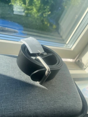 Svart klassiskt bälte i skinn - Snyggt svart bälte i skinn med enkel silverfärgad metallspänne. Perfekt till jeans eller byxor för en clean och stilren look. Bältet har en slät yta och tidlös design som funkar till det mesta.