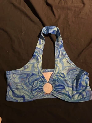 Blå swirl halterneck topp - Säljer en blå halterneck topp med mönster och en snygg ringdetalj framtill. Toppen är ärmlös och croppad